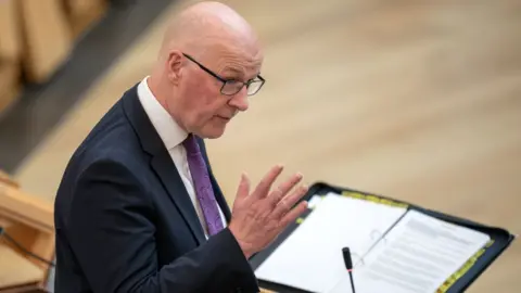 PA Media John Swinney, who is bald with glasses, speaks into a microphone while wearing a business suit 