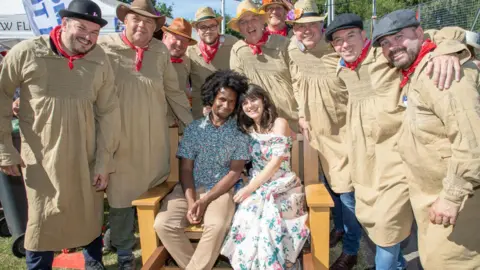 Dunmow Fitch Trials A couple sit on a bench, surrounded by a group of men