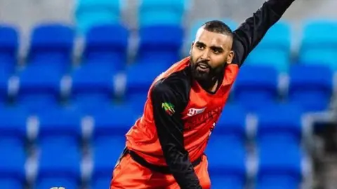 Mohammed Mohammed is pictured in mid-bowl just after releasing the ball. He's wearing a red top with black long sleeves and logos on the chest. His left arm is raised high above his head and his other arm is down low for balance. He looks as if he's exerting himself and concentrating hard. We can tell he's in some sort of sports stadium as the shapes of rows of blue folding seats are visible in the background