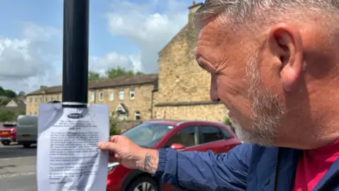 Mark Carter, with short silver hair and a beard, is holding a laminated paper sign attached to a lamp post in front of him.
