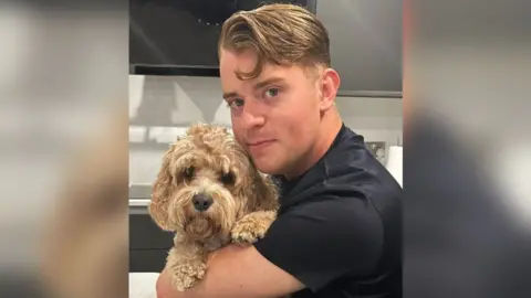 Metropolitan Police Oliver looking at the camera with golden brown short hair, that's longer at the front and wearing a black T-shirt. He is holding a dog golden fluffy dog that is also looking at the camera. 