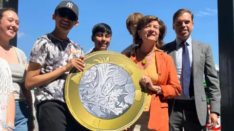 A young male on the left holding a giant £1 coin with Eluned Morgan. Ken Skates, the transport secretary, is stood to the right. Other young people are stood around the pair.