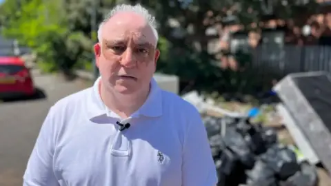 Shafaq Hussain is pictured stood on a road with black bin bags and rubbish piled up behind him. He has white hair and is wearing a white polo T-shirt. He has a small black microphone pinned to his collar. There is a parked red car, pavement and some trees blurred in the background.