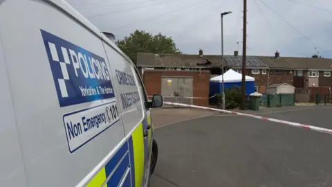 A police investigation scene in a residential neighbourhood.
In the foreground on the left side of the image, a white police van is parked facing away from the camera. The side of the vehicle displays the words "POLICING Yorkshire & The Humber" along with a blue-and-white checkerboard logo. Additional text on the van reads "Crime Scene Investigation" and "Non Emergency 101". Across the middle of the image, red-and-white police tape has been stretched across the road, creating a cordon that restricts access to the area beyond.
In the background, a row of terraced houses stands behind the police cordon. Near the centre of the image, one property shows visible signs of damage, with a large blackened hole in the roof and surrounding scorch marks.
A blue-and-white forensic tent has been erected near the damaged property. 