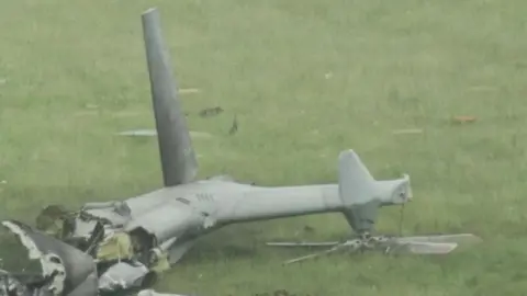 The tail of a grey military helicopter which has crashed in a field in Devon. Wreckage can be seen across parts of the grass.