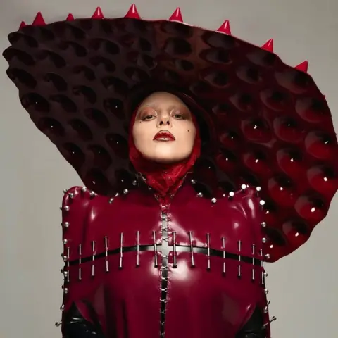 Interscope Lady Gaga mengenakan pakaian kulit plastik merah darah, dijahit bersama dengan kuku, dan atasnya dengan topi berbentuk cakram yang terlalu besar