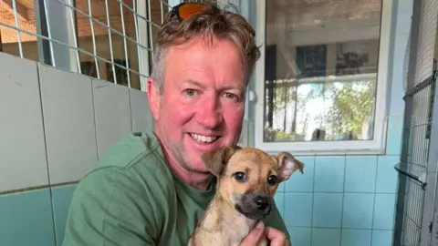 Pen Farthing holding a small dog with light brown fur inside a blue shelter. He is wearing a green T-shirt and smiling.