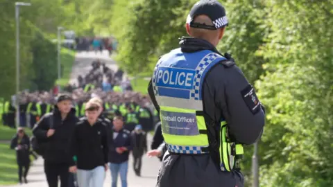 A Devon and Cornwall Police football officer on duty in Central Park near Home Park Stadium in Plymouth. He is in police uniform looking at a large group of supporters and police officers walking towards the ground. There are lots of trees and greenery around him. 