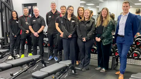 Everyone Active About ten people in black gym uniform are standing and smiling to camera with some rowing machines in the foreground and a bench of weights behind them, reflected in the large mirror on the back wall.
