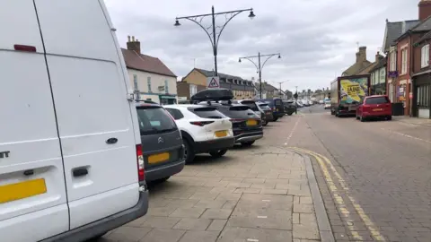 John Devine/BBC Four vehicles, a van and three cars parked on a pavement on the left hand side of a road that has designated bays further along, but they are all full.
There are shops and houses on both sides of the street, and double-yellow lines can be seen on parts of the roadway.