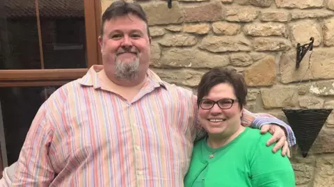 Supplied Steffen and Becky Peddie stand in front of a stone cottage. Steffen, on the left, is wearing a striped pink shirt, has a grey beard and dark hair. He is of a large build. He has his arm around his wife who is quite a lot shorter than him, she also has dark hair, is wearing glasses and has a green twinset on. 