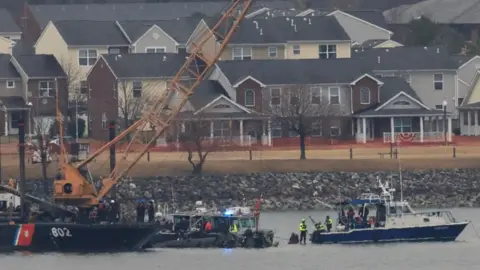 A US Coast Guard vessel with a crane attends the site to try and retrieve the remains of a Black Hawk helicopter near the Ronald Reagan Washington National Airport. 
Homes are seen in the background.