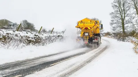 BBC Weather Watchers/Windhater A yellow gritter is driving across a road which is covered in snow.