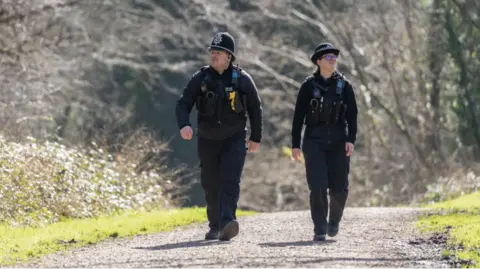 Two police officers, male and female, walking down a footpath. There are trees behind them. One officer is looking to the left and the other is looking to the right.