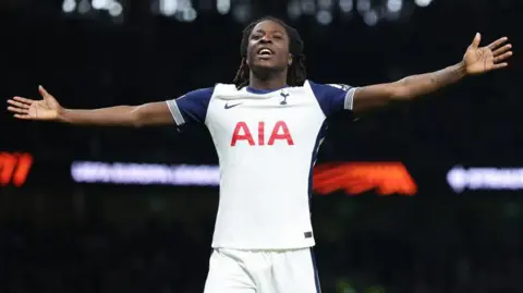 Damola Ajayi stretches his arms out either side in celebration after scoring for Tottenham Hotspur, wearing the club's white home jersey