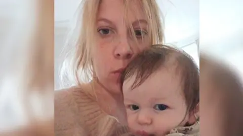 A woman poses for a selfie with her newborn son. She has blonde hair, which is slightly messy, and is wearing a jumper. She is staring at the camera with little expression on her face. The baby boy, who has short brown hair, is also looking at the camera. The woman is holding him close to her face, and looks as though she is giving him a kiss on his forehead.