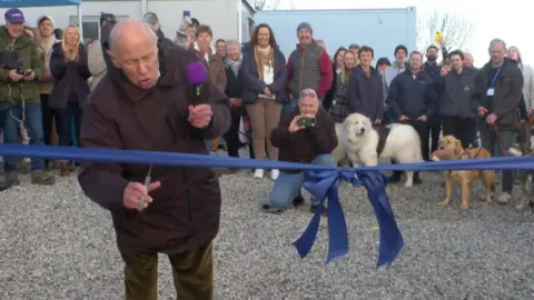 A picture of a man cutting a ribbon. He is holding a microphone and is pictured in front of dogs and people.
