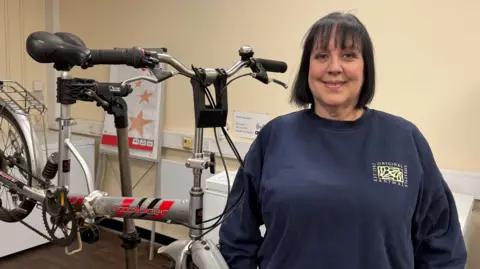 BBC News A lady wearing a navy jumper stands in front of a bike which is being held up by a metal stand. She has black shoulder-length hair with a fringe and is smiling at the camera. The silver bike has a black seat and handlebars.