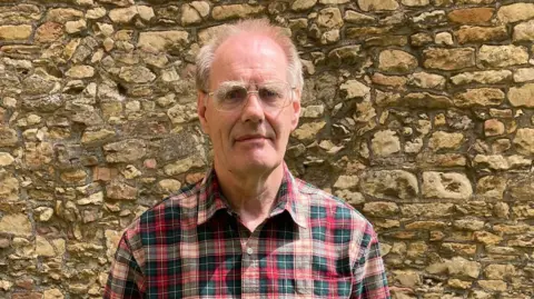 Church warden David Justham standing on a grassed area outside the minster. He is wearing a checked shirt and green trousers and is holding a b walking stick. The wall of the minster can be seen in theackground.