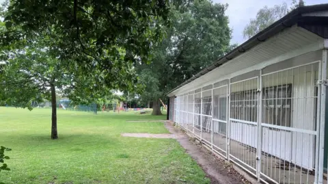 Image shows a recreation ground with green grass and mature trees on the left and a pavilion on the right, covered in white bars.