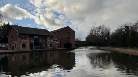 The cafe stands by the side of the canal basin which has a decorative water feature. The building is in shadow and the opposite side of the canal has bare, winter trees.