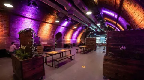 The inside of a bar and events space, with various tables and chairs set up, with the ceiling forming an arch over them 