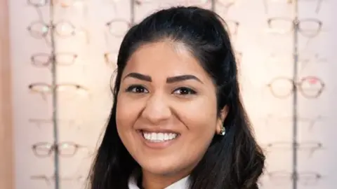 Submitted A portrait picture of Aashika Patel. She has long, black hair scraped back from her forehead. She is smiling at the camera. Behind her, there are four vertical racks with glasses displayed on them. 