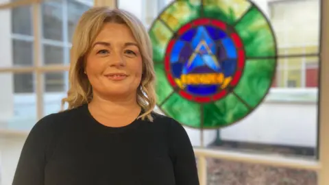 Claire Doherty has long blonde hair. She is wearing a black top and is pictured smiling near a large glass window with a large green, red and blue stained glass motif.