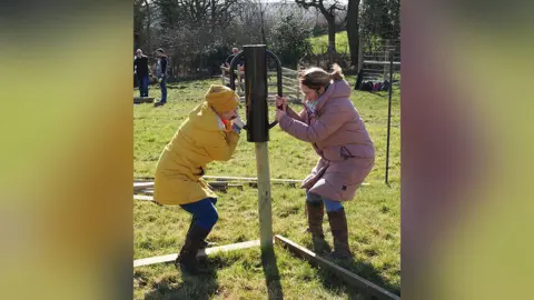 A woman in a yellow coat and hat planting a tree in the ground on farmland along with a woman standing opposite her wearing a pink coat. 
