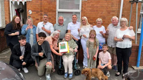 Family photo Phyliss Marshall sat in her wheelchair surrounded by her entire family outside her home in Bridgwater where she was born 100 years ago. She's wearing a green blouse with white spots and beige trousers with a birthday cake being presented to her.