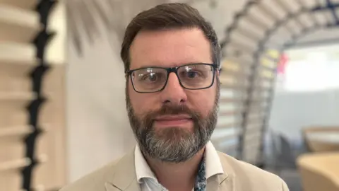 Prof Tristram Hooley from the UCU Union at the University of Derby is looking straight into the camera. He is wearing glasses and a light coloured suit and white shirt.