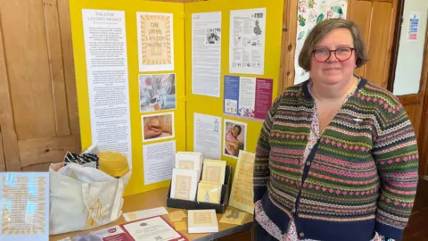 A woman with short dark hair, glasses, a flowery blouse and a stripped knitted cardigan stands beside a trestle table. The table has a large yellow noticeboard on it, numerous leaflets and a cream tote bag filled with a blanket and other items. The wall behind is made of wooden panelling.