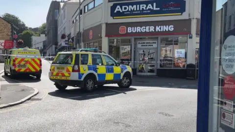 Police vehicles block a town-centre street outside a Burger King. Shops line both sides of the road on a sunny day.