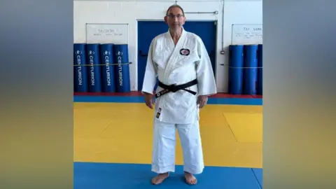 Contributed A man is standing in a judo hall, wearing a judo suit with a black belt. He is barefoot, standing with hands by his side, looking at the camera.