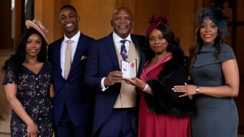 PA Media Devon Malcolm is surrounded by a young woman and man on the left and a woman in a red dress and a second younger woman in a grey dress to the right. The women are all wearing hats and everyone is dress smartly.
