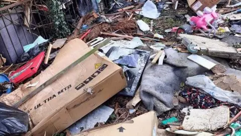 Basildon Council Fly tipped rubbish on the ground outside. There are large cardboard boxes, children's toys, pizza boxes, wood, plastic and what looks to be a broken television and carpet. All have been left in a pile on the ground.