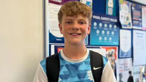 Freddie is wearing a blue patterned sports shirt and a backpack stands in a leisure centre corridor beside a noticeboard covered with posters and pool safety information. A doorway and windows are visible in the background.
