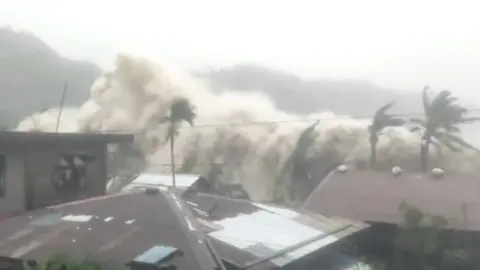 A huge frothy wave crashes over houses and palm trees.