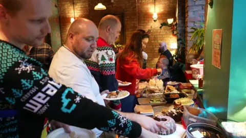 Four people are lined up as they choose cakes from a selection on a table.