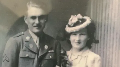 Luke Francis A black and white photograph of a couple. Emlyn is on the left, wearing a hat, with Chester in uniform on the left. They are both smiling at the camera and she is holding on to his arm.