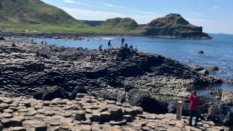 A wide shot of the rocks. People can be seen walking over them. The sea is bright blue in colour and looks calm. The hills in the background are green.