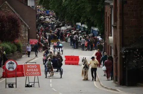 ADAM VAUGHAN/EPA/Shutterstock View of an Appleby street as a several two-wheel horse-drawn carriages ride past onlookers. A number of the carts are parked on the right-hand side of the image at the bottom of a hill. Barriers and road signs tell people the road ahead is closed to traffic.