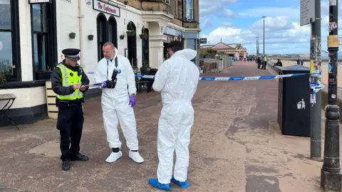 BBC A police photographer, wearing a white hazmat suit chats to a uniformed officer carrying a clipboard as another office, also wearing a hazmat suit, looks on. The beach promenade in the background is cordoned off with blue and white police tape