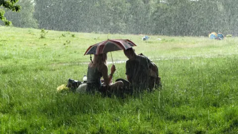 Getty Images Two people sat on grass under an umbrella. Their backs are to the camera.