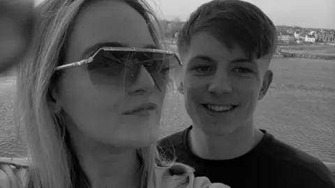 A black and white photo of the teenage couple. McKenzie, smiling, is clean-shaven with a side-swept fringe haircut with faded back and sides. Olivia, taking the photo, wears sunglasses. Behind them is a body of water - which looks like the sea - and houses can be seen on a shoreline further away.