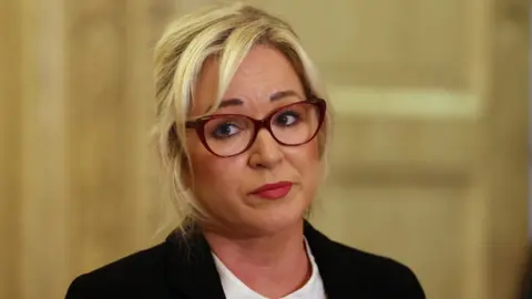 Michelle O'neill - a woman with blonde hair, red glasses and a black blazer
