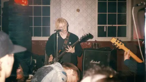 Historic England/Home of Metal A man with short blond dreadlocks plays the guitar in a pub, with members of the audience visible at the bottom of the photo