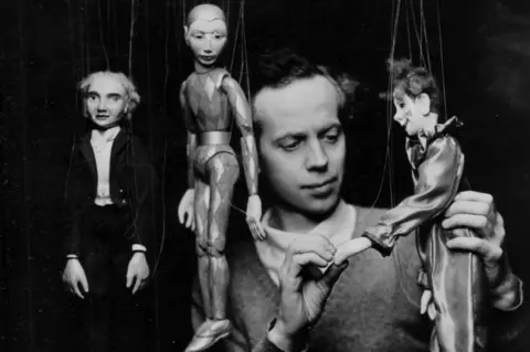 A black and white photograph of a man with three marionettes suspended by string. He is supporting one of them with his hand.