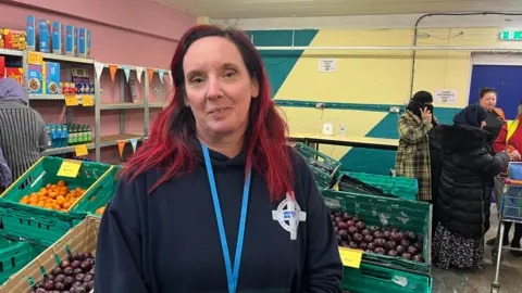 BBC Julie-Anne Webb standing in front of a surplus food shop at St Vincent's. Fruit is pictured in crates and some cereal boxes and drinks are on shelves to the left. People are shopping in the background.