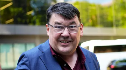 Graham Linehan photographed outside Southwark Crown Court in a blue suit and purples shirt, he is wearing glasses